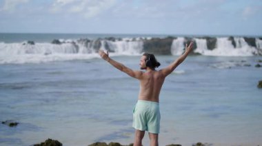 Genç adam, kulaklıkları açık, kolları açık, neşeli bir şekilde okyanusa bakıyor, çarpıcı dalgaların ortasında coşkusunu ve özgürlüğünü ifade ediyor.