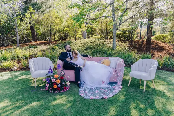 Bir çift, renkli çiçeklerle çevrili, canlı bir bahçede, eski bir kanepede huzurlu bir anın tadını çıkarıyor. Düğün kutlamalarının cazibesini arttırıyor ve neşeli bir zemin oluşturuyor.