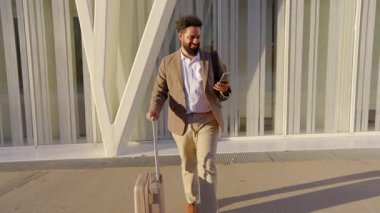 Şık ve modern Afro-Amerikan iş adamı bavuluyla güvenle yürüyor, çağdaş bir seyahat estetiğini somutlaştırıyor, bir sonraki önemli randevusuna ya da iş toplantısına tamamen hazır.