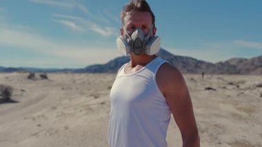 Uçsuz bucaksız, çorak ve çorak bir çöl manzarasında, yalnız bir adam hayatta kalma ve izolasyon temalarını anımsatan bir maske takıyor.