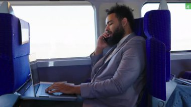 Trendeki Afrikalı Amerikalı işadamı telefonda konuşurken dizüstü bilgisayarını kullanıyor. Kusursuz bir şekilde giyinmiş, profesyonellik yayıyor ve modern iş seyahatlerini ve çoklu görevleri somutlaştırıyor.