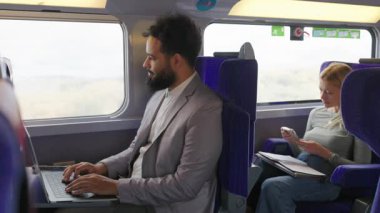 Bir iş arkadaşı okurken Afrikalı Amerikalı bir işadamı tren masasında dizüstü bilgisayarını kullanıyor. Bu sahne, modern iş seyahatlerini ve mesleki yaşamlarını yakalıyor, odaklanma ve motivasyonlarını gösteriyor.
