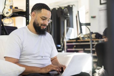 A bearded man sits in a bright, contemporary workspace, comfortably focused on his laptop, immersing himself in creative tasks and exuding both inspiration and concentration while working clipart