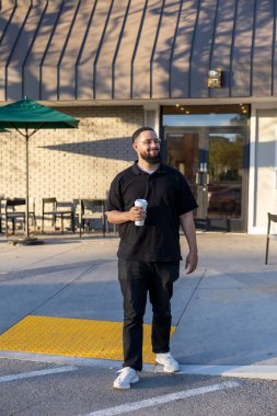 Siyah gömlekli neşeli bir adam elinde kahve fincanıyla kafeden çıkıp güneşli havanın tadını çıkarıyor.