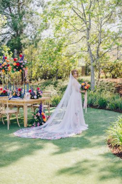 Göz kamaştırıcı bir bahçede zarif bir gelin, akışkan bir duvağı olan büyüleyici bir gelinlik giyiyor, renkli çiçeklerle çevrili, ve güzelliği arttıran zarif bir masa.