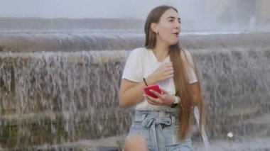 Seyahat ederken akıllı telefonunu kullanan genç bir bayanın portresi. Arka planda şehir çeşmesi olan neşeli ve tasasız görünen genç bir kızın yakın çekimi. Yüksek kalite 4k görüntü