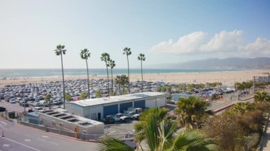İskeledeki Meşhur Lunapark 'ın yanında el kamerasıyla çekilmiş bir fotoğraf. Santa Monica plajı, Kaliforniya. Mavi parlak gökyüzü güneşli bir günde. Arabalar park yerinde. Avuçlar ve üçler. Yüksek kalite 4k görüntü