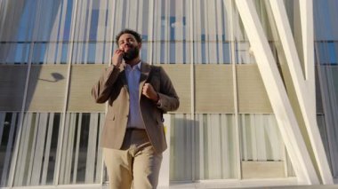 Bir iş adamı modern bir binanın dışında yürüyor, bir telefon görüşmesine katılıyor. Şık bir takım elbise giymiş, kendine güveni tamdır. Başarıya ve liderliğe olan bağlılığını yansıtır.