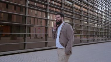 Başarılı iş adamı, telefonuyla bir konuşma yaparken, hareketli, gösterişli, modern şehir ortamında güvenli adımlar atar, profesyonel tavrını ve tarzını sergiler.