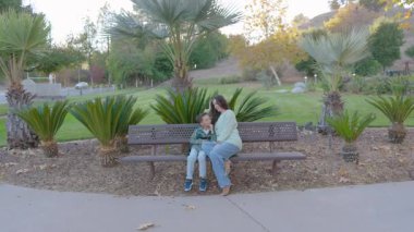 Yürek ısıtan bir sahnede, sevgi dolu bir anne ve tatlı çocuğu birlikte huzurlu bir anın tadını çıkarırken, aynı zamanda tamamen canlı, yemyeşil bir yeşil bankta oturuyorlar.