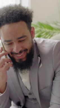Hafif takım elbiseli gülümseyen Afrikalı Amerikalı işadamı şık bir çalışma alanında rahat bir şekilde oturuyor, yemyeşil bitkiler ve parlak pencereler arasında bir telefon görüşmesi yapıyor.