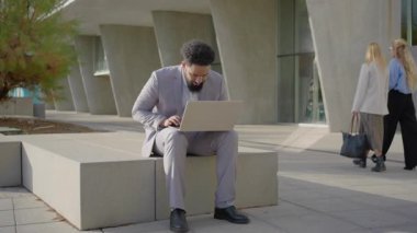 Canlı bir şehir ortamında, Afro-Amerikan bir işadamı özenle dizüstü bilgisayarı üzerinde çalışıyor, şık, modern bir bankta oturuyor, bir kadın ise hareketli şehir atmosferine katkıda bulunarak geziniyor.