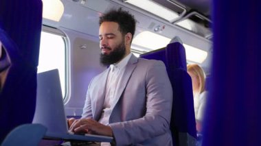 Şık takım elbiseli başarılı bir Afro-Amerikan işadamı, trende rahat bir şekilde otururken dizüstü bilgisayarına odaklanır, profesyonelliği ve yüksek verimliliği somutlaştırır.