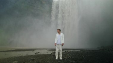 Genç adam kendine güvenen beyaz elbisesiyle ikonik Skogafoss şelalesinin yanında duruyor. Etrafı dönen sisle çevrili ve manzaralı İzlanda 'nın nefes kesici güzelliğiyle çevrili.