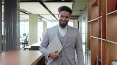 Tarz sahibi Afro-Amerikan işadamı gülümseyerek ve dizüstü bilgisayarını dinamik bir ofis ortamında, karizmada ve kurumsal dünyada başarının özünde tutarak güven ve profesyonellik yayıyor.