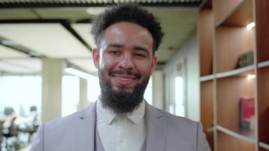 Gülümseyen bir Afro-Amerikan işadamı, işbirliği ve yaratıcılığı teşvik eden, profesyonellik ve liderlik havası yayan, stil sahibi, modern bir çalışma alanında kendine güvenerek duruyor.