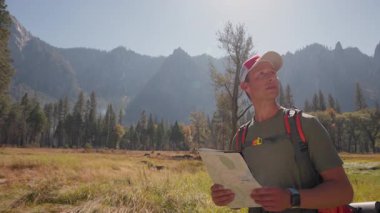 Yosemite Milli Parkı 'nda unutulmaz bir maceraya çıkmış genç beyaz bir erkek, ayrıntılı bir harita kullanarak, şapka takıyor ve seyahat sırt çantası takıyor, ikonik tepelerle dolu büyüleyici manzaralarda geziniyor.