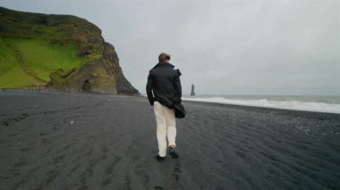 Genç adam İzlanda 'daki Reynisfjara' nın siyah kumlu plajı boyunca sakin bir şekilde yürüyor. Burası Güney İzlanda 'nın çarpıcı bölgesinde nefes kesici sahil kayalıklarıyla güzel bir şekilde çevrili.