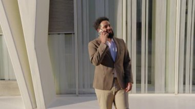 Afro-Amerikalı bir işadamı, şehir merkezinde şık bir takım elbise giymiş ve etkileyici bir modern mimariyle çevrelenmiş bir şekilde seyahat ederken aktif olarak bir telefon görüşmesi yapmaktadır.