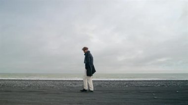 Genç adam, Reynisfjara Sahili 'nin ikonik, çarpıcı güzellikte, rüzgarlı, siyah kumsalında, İzlanda' nın pitoresk güney kıyısında, güzelliklerle çevrili, sakin bir şekilde geziniyor.