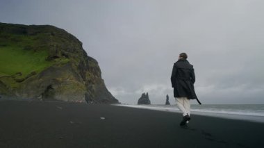 Genç bir adam, İzlanda 'da Reynisfjara' nın eşsiz siyah kumlu plajında duruyor, rüzgarı kucaklıyor ve onu çevreleyen büyüleyici manzaranın nefes kesici güzelliğini içine çekiyor.