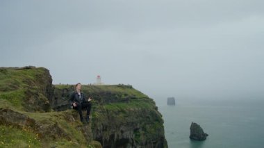 Dyrholaey Deniz feneri yakınlarındaki Sersemletici kayalıklarda oturan adam İzlanda 'nın engebeli kıyı şeridine nefes kesici manzaralar sunuyor, sisli bir zemin üzerine kurulmuş, dış dünyevi bir keşif ve macera çağrıştırıyor.