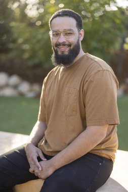 Kahverengi bir gömlek giymiş, hayat saçan canlı ve yemyeşil bir ortamda rahat bir şekilde oturan, bakımlı sakallı, neşeli bir Afrikalı Amerikalı adamın detaylı portresi.