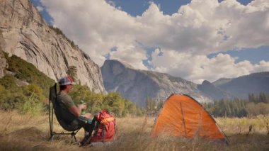 Bir adam dinlenmek için arkasına yaslanır ve nefes kesici Yosemite Milli Parkı 'ndaki çadırında dinlenir. Etkileyici dağlarla çevrili ve el değmemiş vahşi doğanın olağanüstü güzelliğiyle çevrelenmiş.