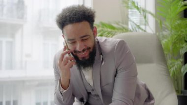 Hafif takım elbiseli gülümseyen Afrikalı Amerikalı işadamı şık bir çalışma alanında rahat bir şekilde oturuyor, yemyeşil bitkiler ve parlak pencereler arasında bir telefon görüşmesi yapıyor.