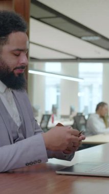 Hafif takım elbiseli bir Afrikalı Amerikalı laptopuna odaklanır. Çevresi çeşitli işlerde çalışan profesyonellerle çevrilidir. Dinamik bir atmosfere katkıda bulunur.