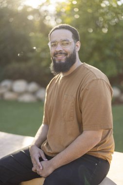 Neşeli bir adam, bakımlı bir sakalı olan, dışarıda rahat bir şekilde oturmakta, sıcaklığı, pozitifliği ve neşeli bir atmosferi olan, canlı, doğal bir ortam ile çevrili.