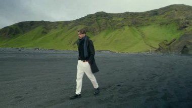 Tarz sahibi adam Reynisfjaras 'ın koyu siyah kumsalında duruyor, İzlanda' nın güney kıyısındaki canlı yeşil uçurumların güzelliğine dalmış, çevresini keşfediyor ve huzurlu atmosferin tadını çıkarıyor.