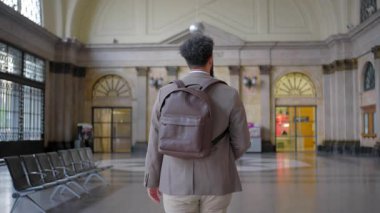 Bir tren istasyonunun hareketli ortamında, profesyonel görünümlü bir işadamı bir elinde şık bir sırt çantası taşırken özgüvenle, zahmetsizce bir akıllı telefonu dengede tutar.