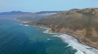 Havadan çekilen görüntüler, 1. Otoyol 'daki çarpıcı Big Sur sahili boyunca güçlü okyanus dalgalarıyla harmanlanmış canlı günbatımı renkleri sergiliyor, Kaliforniya kıyılarının eşsiz cazibesini vurguluyor.