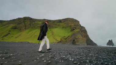 Reynisfjara 'nın çarpıcı siyah kumsalında yürüyen genç adamın tüm vücudu dramatik uçurumlar ve İzlanda Güney Sahili' nin nefes kesici manzaralarıyla çevrili.