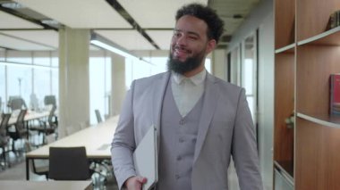 Afro-Amerikalı bir işadamı, şık giyinmiş, kendine güvenen, modern ve parlak bir ofis alanında yürüyor, güçlü bir profesyonellik ve hırs hissi yayıyor.