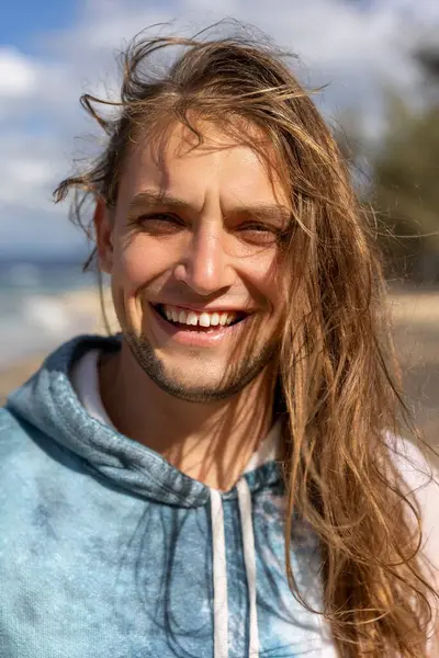 Uzun dalgalı saçları olan neşeli genç bir adam sahilde duruyor, büyüleyici okyanus zeminine karşı kaygısız neşenin özünü mükemmel bir şekilde yansıtan parlak bir gülümsemeyle ışıldıyor.
