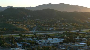 İnsansız hava aracı görüntülerinde Los Angeles 'ın canlı banliyöleri, verimli tepeler, görkemli dağlar ve hareketli otobanlar, manzarayı güzelleştiren çarpıcı bir günbatımı arka planına karşı gösteriliyor.