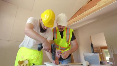 İnşaat alanında, gerekli güvenlik donanımlarını giymiş iki dalgıç inşaat işçisi, tamamen görevlerine odaklanmış durumdalar, tahta desteğini vidalıyorlar, takım çalışmasının özü ve sıkı çalışma.
