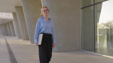 Bir iş kadını, elinde bir dizüstü bilgisayarla modern bir mimari yolda güvenli adımlar atıyor. Kıyafeti profesyonel ama yaklaşılabilir bir tavrı yansıtıyor. İş yeri için mükemmel..