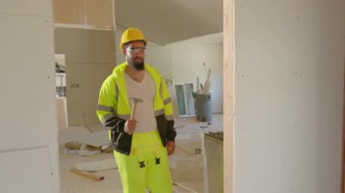 Odaklanmış gülümseyen Afro-Amerikan inşaat işçisi. Parlak güvenlik giysileri içinde. Bitmemiş bir binada çalışırken elinde ustaca bir çekiç tutuyor. İnşaat, güvenlik protokolleri, profesyonellik içeriği