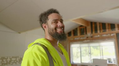 Parlak sarı güvenlik yeleği giyen neşeli siyah ve hevesli inşaat işçisi kısmen yenilenmiş bir binanın içinde duruyor. Pozitif, bağlılık ve profesyonellik yayıyor.