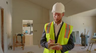 Güvenlik kaskı ve yansıtıcı yelek giyen profesyonel mühendis, yoğun bir inşaat alanını teftiş ediyor, her şeyin yolunda gittiğinden emin olmak için bir not defteriyle not alıyor.