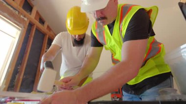 Parlak kasklı ve yansıtıcı yelekli iki yetenekli inşaat işçisi, ev yenileme, istisnai takım çalışması ve iletişimi gösterirken profesyonel olarak çeşitli araçlar kullanarak işbirliği yapıyorlar.