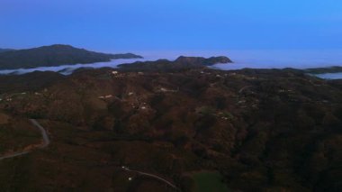 Malibu dağlarının sersemletici hava aracı görüntüleri canlı yeşil tepeler, dönen sis ve gün batımında yumuşak bulutlar sergiliyor. Açık hava macerası için ideal bir ortam yaratıyor.