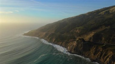 Gün batımında Big Surs 'ın kıyılarında hava fotoğrafları sergileniyor. Sıcak renkler Pasifik' e yansıyor. Gezginler 1. Otoyol 'da manzaralı bir yolculuğun keyfini çıkarırken unutulmaz bir doğa deneyimi yaratıyor.