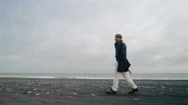 Reynisfjaras 'ın eşsiz jeolojisi ve kıyı manzarası için İzlanda' daki kara kumsalda yürüyen şık bir adam dramatik bir gökyüzü altında, dalgalar çarpıyor, barışçıl bir atmosfer yaratıyor.