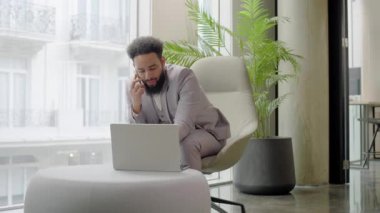İyi giyimli, şık takım elbiseli, Afro-Amerikan profesyonel iş adamı modern ofis alanında rahat bir şekilde oturduğu bir telefon görüşmesiyle meşgul.