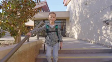 Okul binasının dışındaki merdivenlerden mutlu bir şekilde iniyor. Renkli bir ekose gömlek giymiş ve okul çantasını taşıyor. Güneşli bir günde, sıcak bir ortamda eğleniyor.