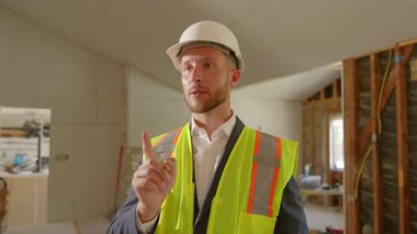 Güvenlik teçhizatı içinde bir inşaat yöneticisi elinde bir defter tutarken şantiyedeki işleri sayıyor. Tamamlanmamış iç mekan, sürmekte olan yenilemeleri ve devam etmekte olan çalışmaları vurguluyor